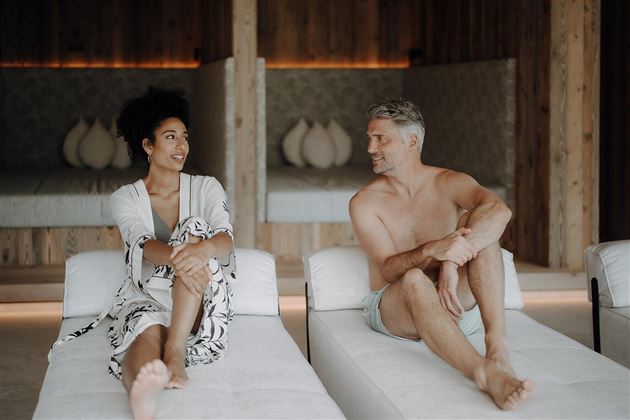 A couple sits relaxed on loungers in a stylish room. They smile at each other and enjoy their time together.