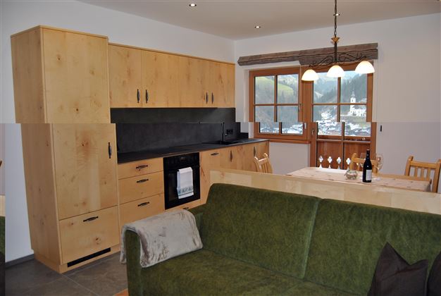 A modern kitchen with wooden cabinets and a dining table. In the foreground, a cozy sofa is visible.