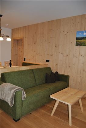 A modern living room with a green sofa and a wooden table. The walls are made of light wood and there is a small picture on the wall.