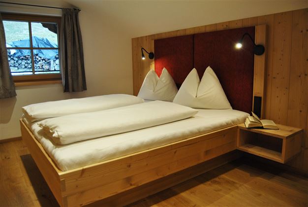 A cozy bedroom with a double bed and wooden details. The walls are light, and there is a window with curtains.