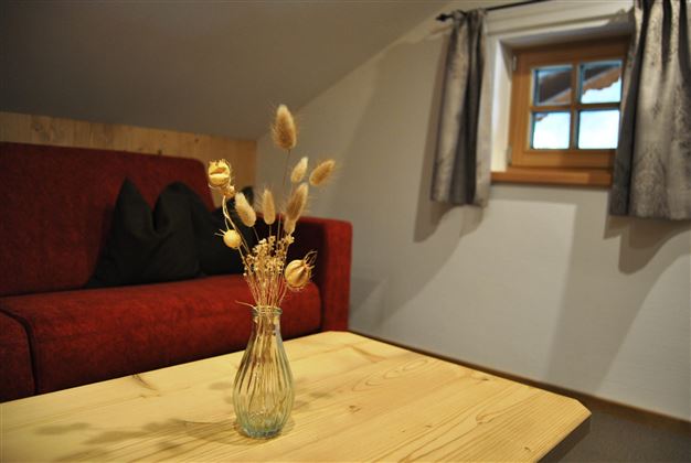 A cozy room with a red sofa and a small wooden table. On the table, there is a vase with dried flowers, and a window lets in natural light.