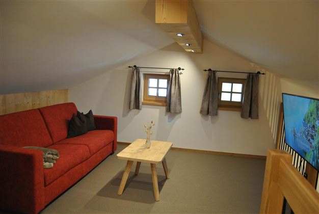 A cozy living space with a red sofa and a wooden table. Two windows provide a bright atmosphere.