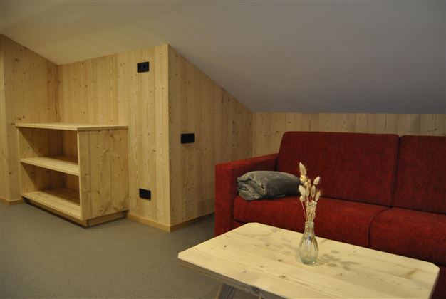 A cozy room with a red sofa and a wooden table. The walls are made of wood, creating a warm atmosphere.