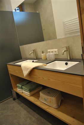 A modern bathroom with a wooden washbasin and two sinks. The walls are painted in neutral colors, and a towel is placed next to the sinks.