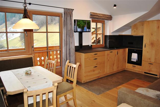 A cozy modern kitchen with wooden furniture and a dining area. Large windows offer a view of the surrounding landscape.