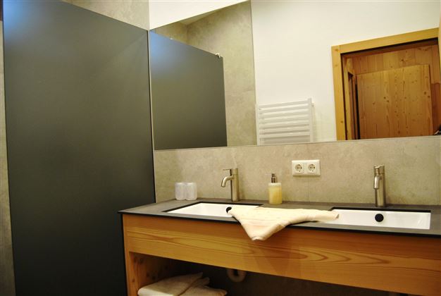 A modern bathroom with two sinks and a wooden shelf. The walls are designed in neutral tones and there is a dressing wall for privacy.