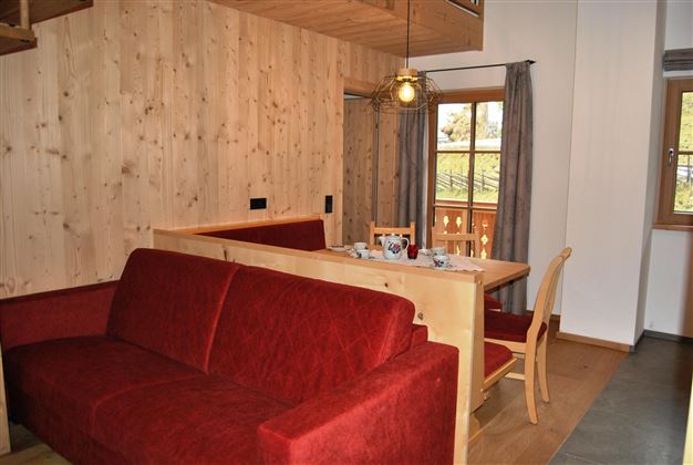 A cozy living area with a red sofa and a wooden table. Large windows let in plenty of light and offer a view of nature.