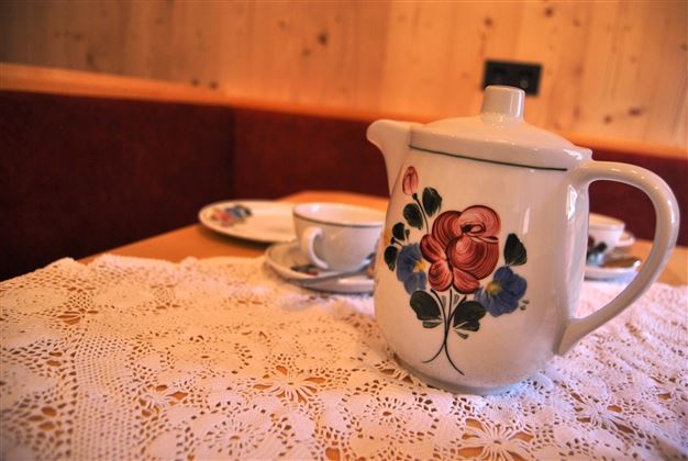 An elegant teapot with a floral design is on a table. In the background, tableware and a tablecloth can be seen.