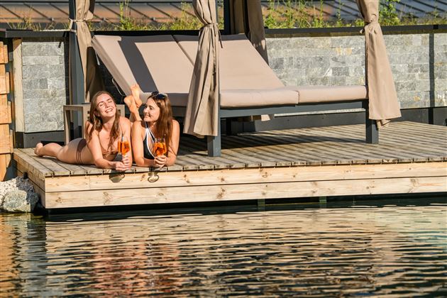 Zwei Frauen liegen entspannt auf einer Holzplattform am Wasser und genießen Cocktails. Im Hintergrund ist ein Schattenplatz mit Liegestühlen zu sehen.