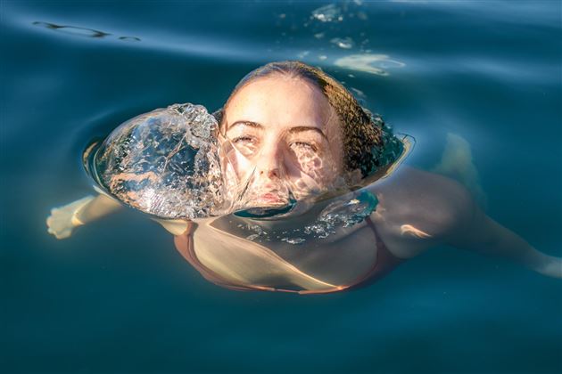 Ein Mädchen schwimmt unter Wasser und hat eine Luftblase um ihr Gesicht. Das Wasser ist klar und reflektiert das Licht.