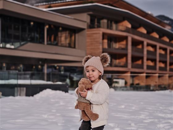 Ein kleines Kind steht im Schnee und hält ein Teddybär. Im Hintergrund ist ein modernes Gebäude zu sehen.
