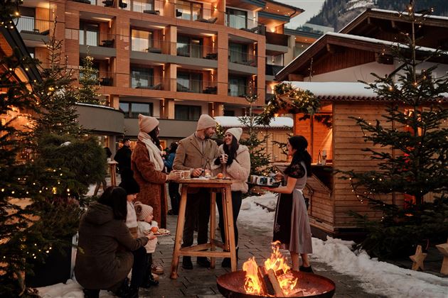 Eine Gruppe von Menschen steht um einen Tisch in einem winterlichen Außenbereich. Im Hintergrund sieht man ein Holzhaus und Weihnachtslichter, während ein Feuer knistert.