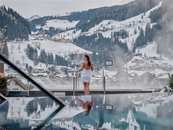 Eine Frau in einem Bademantel steht am Rand eines Pools und blickt auf schneebedeckte Berge. Der Pool spiegelt die Landschaft wider und schafft eine entspannende Atmosphäre.