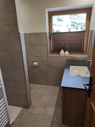 A modern bathroom with tiles and a window. There is a hand wash basin and a privacy screen.