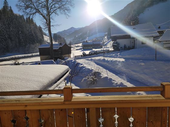 A snowy landscape with hilly mountains in the background and a sun illuminating the sky. In the foreground, snow-covered rooftops and a balcony can be seen.