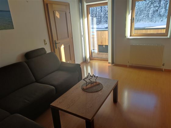 A cozy living room with a gray couch and a wooden table. Large windows offer a view of the snow-covered landscape.