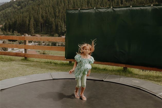 Ein Mädchen springt fröhlich auf einem Trampolin. Im Hintergrund sind Bäume und eine grüne Wand zu sehen.