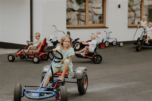 Eine Gruppe von Kindern fährt auf Pedal-Gokarts. Die Szenen spielen sich auf einem gepflasterten Bereich vor einem Gebäude ab.