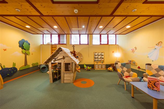 A cozy playroom with a wooden playhouse and colorful wall murals. There are comfortable armchairs and toys on the floor.