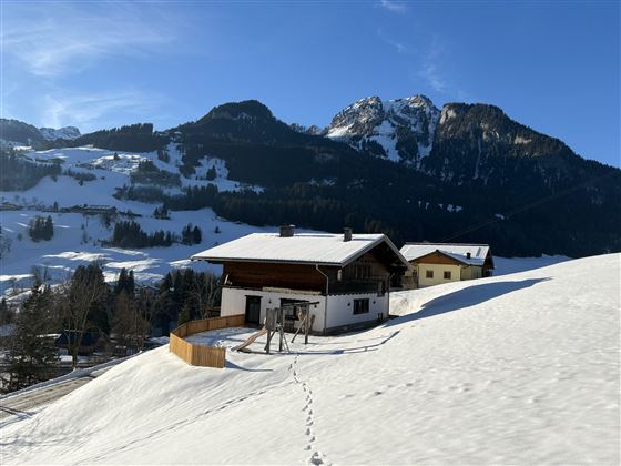 Ein verschneites Landschaftsbild mit einem traditionellen Chalet im Vordergrund. Im Hintergrund erheben sich majestätische Berge unter einem klaren Himmel.
