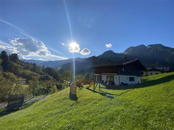 Ein gemütliches Haus auf einer Wiese, umgeben von Bergen und einem klaren blauen Himmel. Die Sonne scheint und der Blick auf die Natur ist malerisch.