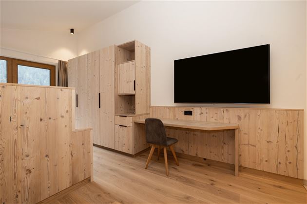 Ein modernes Arbeitszimmer mit einer Holzoptik. Es gibt einen Fernseher an der Wand und einen Schreibtisch mit einem Stuhl.