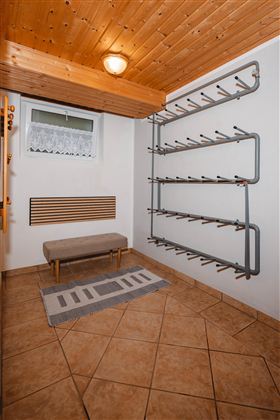 A hallway with a brown tiled floor and a wooden ceiling.  
On the wall, there is a shoe rack and a bench.