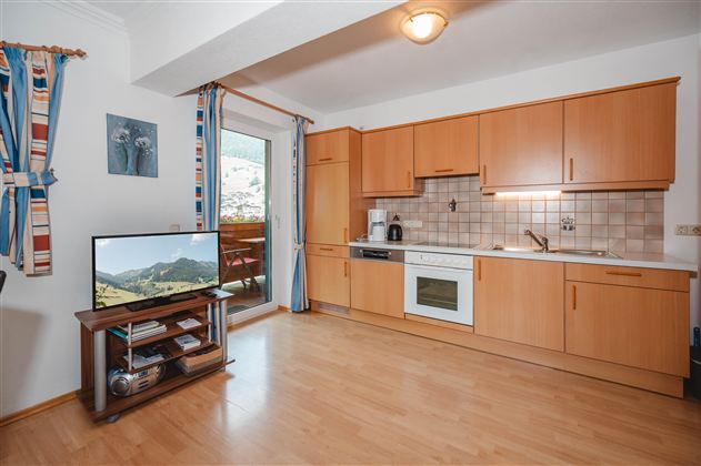 A modern kitchen with wooden cabinets and a light floor. In the room, there is a television and there is a view of the mountains through the window.