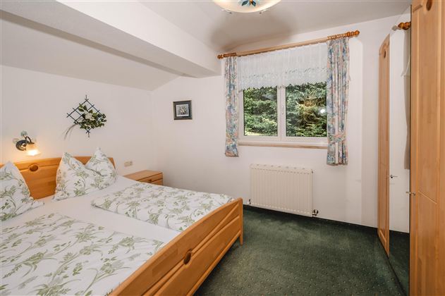 A cozy bedroom with two beds and green bedding. The windows offer a view of nature and the room is bright and pleasantly furnished.