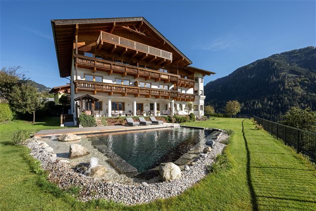 Ein schönes, modernes Haus mit einem großzügigen Garten und einem Teich. Im Hintergrund sind grüne Berge und klarer Himmel zu sehen.