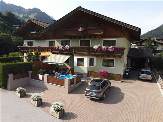 Ein gemütliches Haus mit Blumen auf den Balkonen und einer Einfahrt. Im Vordergrund sind Menschen, die sich unter einem Sonnenschirm entspannen.