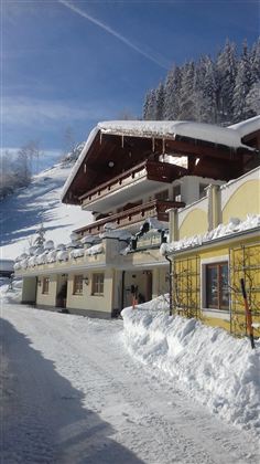 A charming, snow-covered chalet in the mountains. The sky is clear and blue, surrounded by snowy trees.