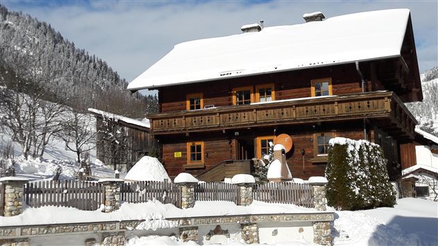 A beautiful wooden house in the snow with a balcony. The surroundings are wintry, surrounded by mountains and snow-covered trees.