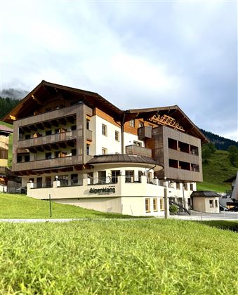 Ein modernes Hotel mit Holzfassade, umgeben von grünen Wiesen und Bergen. Der Himmel ist teilweise bewölkt, was eine ruhige Atmosphäre schafft.