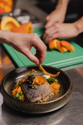 Ein kunstvoll angerichteter Teller mit Fisch, Gemüse und Kräutern. Im Hintergrund wird zusätzlich Obst vorbereitet.