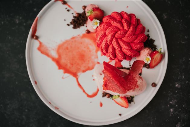 Ein kunstvoll angerichtetes Dessert mit frischen Erdbeeren und einer cremigen Textur. Auf dem Teller sind Schokoladenstückchen und eine fruchtige Sauce zu sehen.