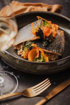 Ein kunstvoll angerichteter Teller mit gebratenem Fisch und buntem Gemüse. Im Hintergrund ist ein Glas Wein und Besteck sichtbar.