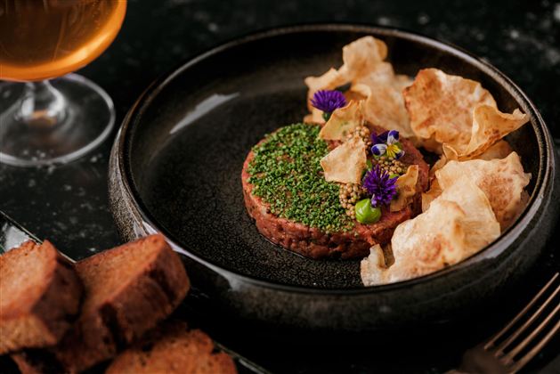 Ein appetitlicher Teller mit Tatar, garniert mit bunten Kräutern und essbaren Blüten. Dazu knusprige Chips und Scheiben von dunklem Brot.