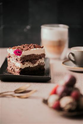 Ein Stück Schokoladenkuchen auf einem schwarzen Teller, garniert mit einer Blume. Im Hintergrund sind eine Tasse Kaffee und ein Glas Getränk zu sehen.