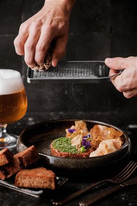 Eine Person reibt eine Zutat über ein dekoratives Gericht in einer Schüssel. Im Hintergrund steht ein Glas Bier und daneben liegen einige Stücke Brot.