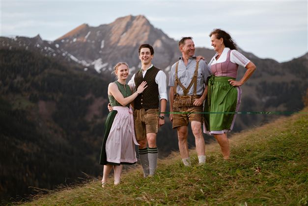 Eine Gruppe von vier Personen in traditioneller Tracht steht auf einer Wiese mit Bergen im Hintergrund. Sie lächeln und wirken fröhlich in der Natur.