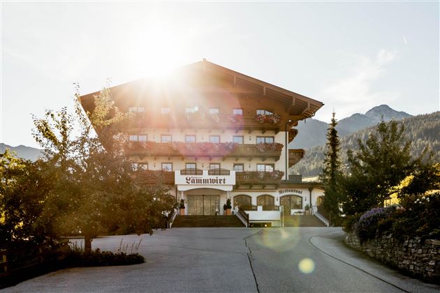 Ein malerisches Hotelgebäude umgeben von Bäumen und Bergen. Die Sonne scheint warm auf die Fassade und schafft eine einladende Atmosphäre.