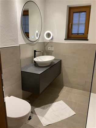 Ein modernes Badezimmer mit einer eleganten Waschbeckenablage und einem runden Spiegel. Die Wände sind hell und die Fenster sorgen für angenehmes Tageslicht.