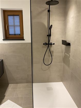 Ein modernes Badezimmer mit einer begehbaren Dusche. Die Wände sind in neutralen Farbtönen gefliest, und ein Fenster lässt Tageslicht herein.