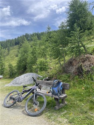 Ein Mountainbike liegt neben einer Bank im Grünen. Im Hintergrund sind Bäume und sanfte Hügel zu sehen.