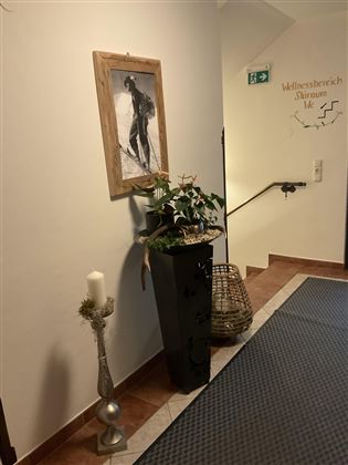 Ein Flur mit einem Gemälde an der Wand und einer stilvollen Dekoration. Auf der linken Seite steht eine Pflanze in einem Behälter und eine mit einem Kerzenhalter verzierte Säule.