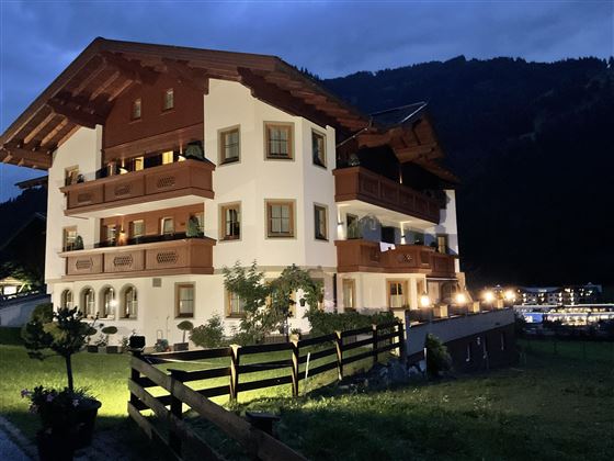 Ein gemütliches Alpenhaus mit mehreren Balkonen, beleuchtet in der Dämmerung. Umgeben von Wiesen und Bergen, strahlt es eine charmante Atmosphäre aus.
