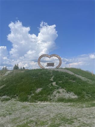 Eine grüne Hügelkuppe mit einer Herzschaukel vor einem blauen Himmel. Die Wolken sind hell und sorgen für eine freundliche Atmosphäre.
