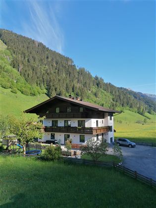 A picturesque house surrounded by green meadows and mountains. The sky is clear and blue.