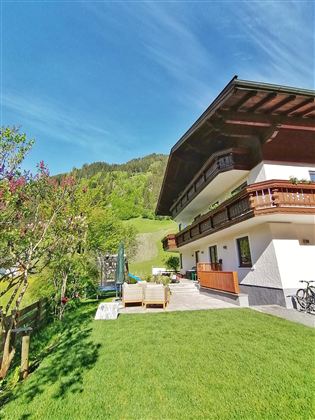 A modern house with a beautiful garden. In the background, green hills and blue sky can be seen.